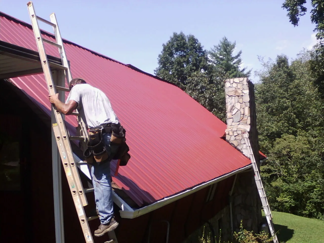 Roofer on a ladder beside a red metal roof and stone chimney — Soffit & Fascia Repair by Roofing Brothers in Millerton